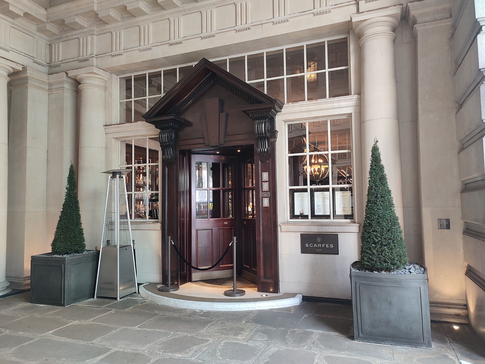 The entrance in the courtyard of the Rosewood London, under a wooden porch between clipped trees.