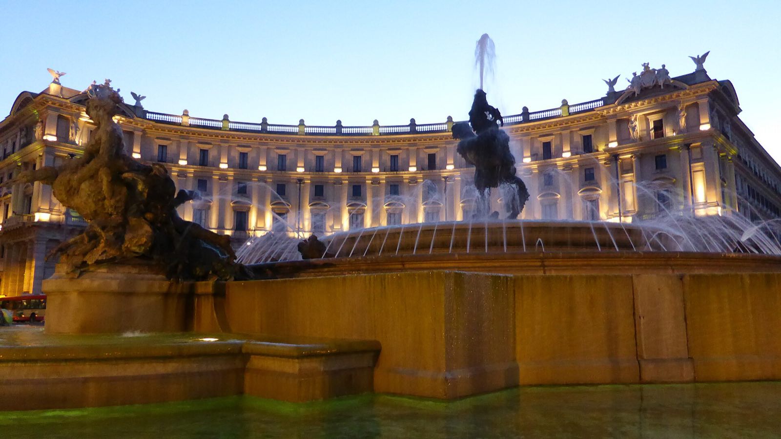 The curved palace of the hotel and the Fountain of the Naiads on Piazza della Repubblica at dusk.