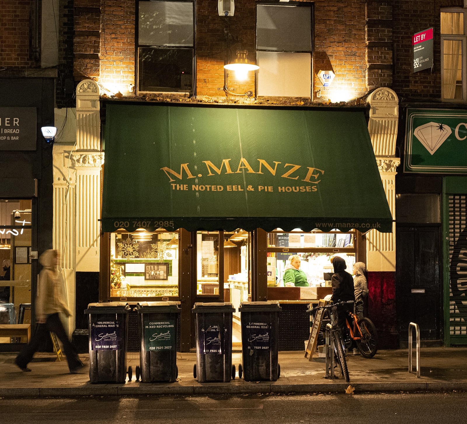The front of M.Manze lit at night, with its name on a green and gold fascia.