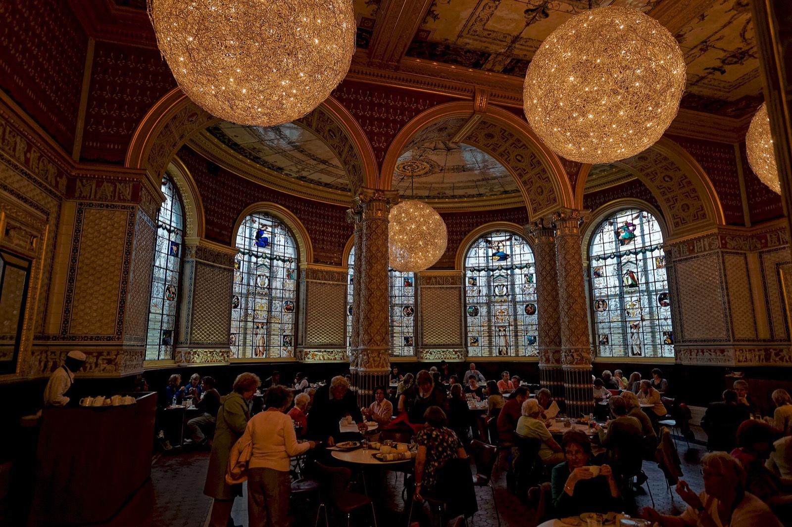 The Gamble Room of the V&A Café, tiled columns and arches under great round lamps, with visitors at the tables.