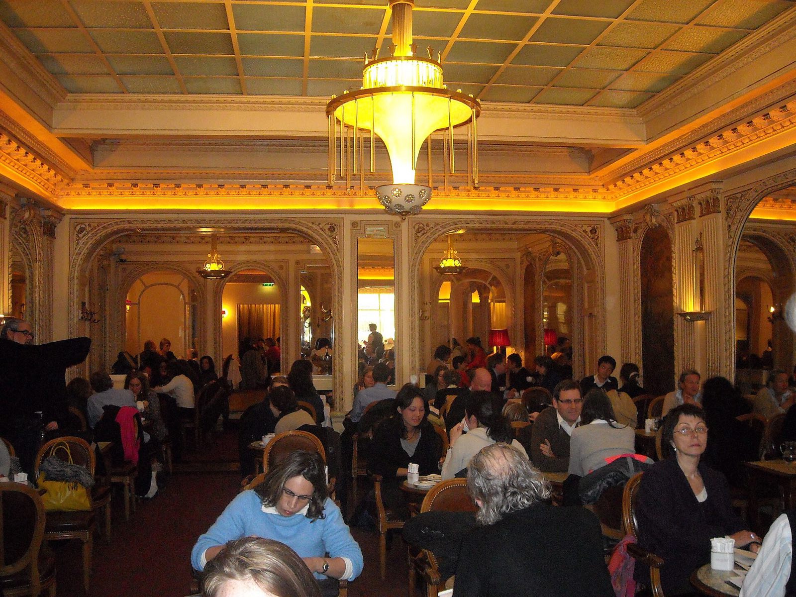 The tea room of Angelina, full of people at small tables under gilded mouldings and lamps.