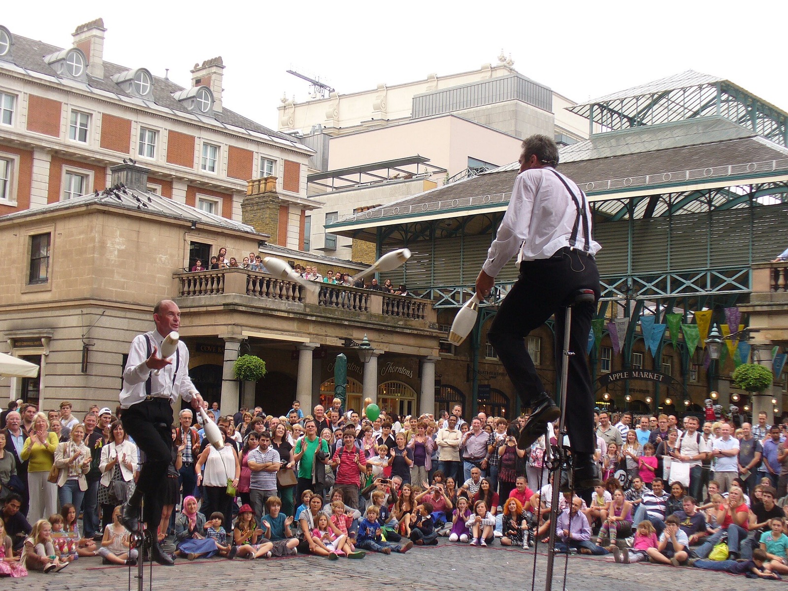 A street performer on a tall unicycle in front of a crowd on the Covent Garden piazza.