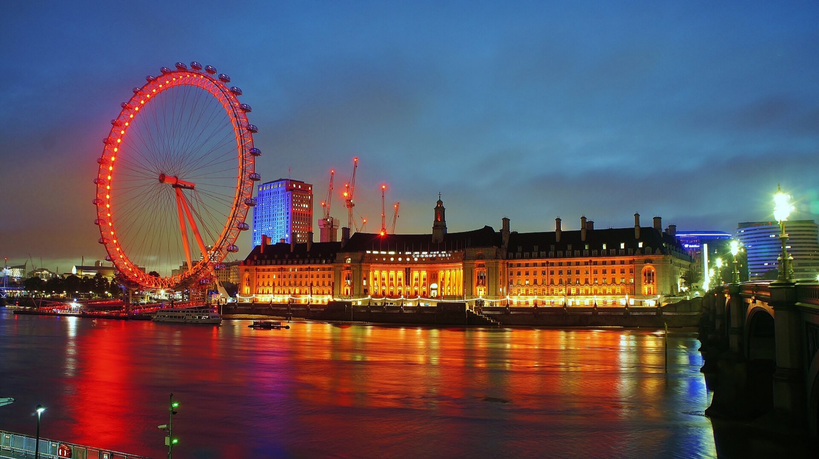 The London Eye lit red at night beside County Hall, reflected in the river.