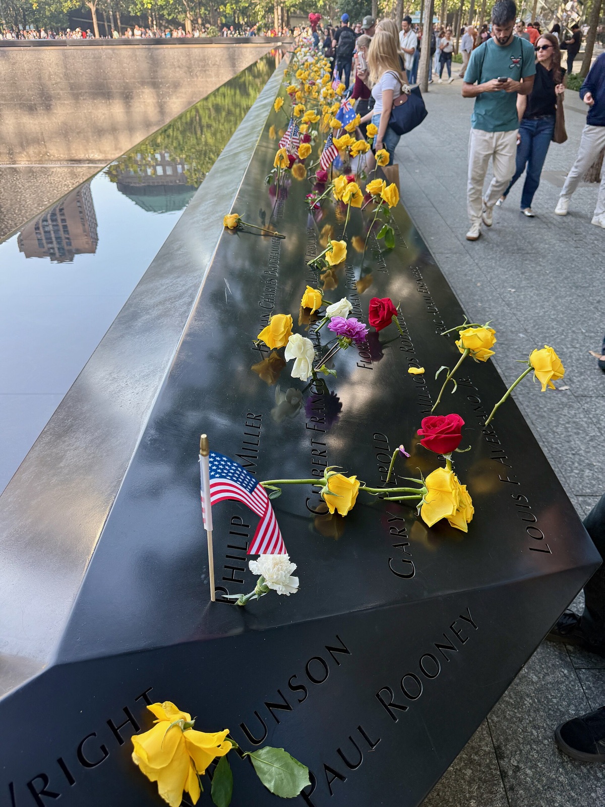 Flowers and small flags left in the names cut into the bronze edge of a memorial pool.