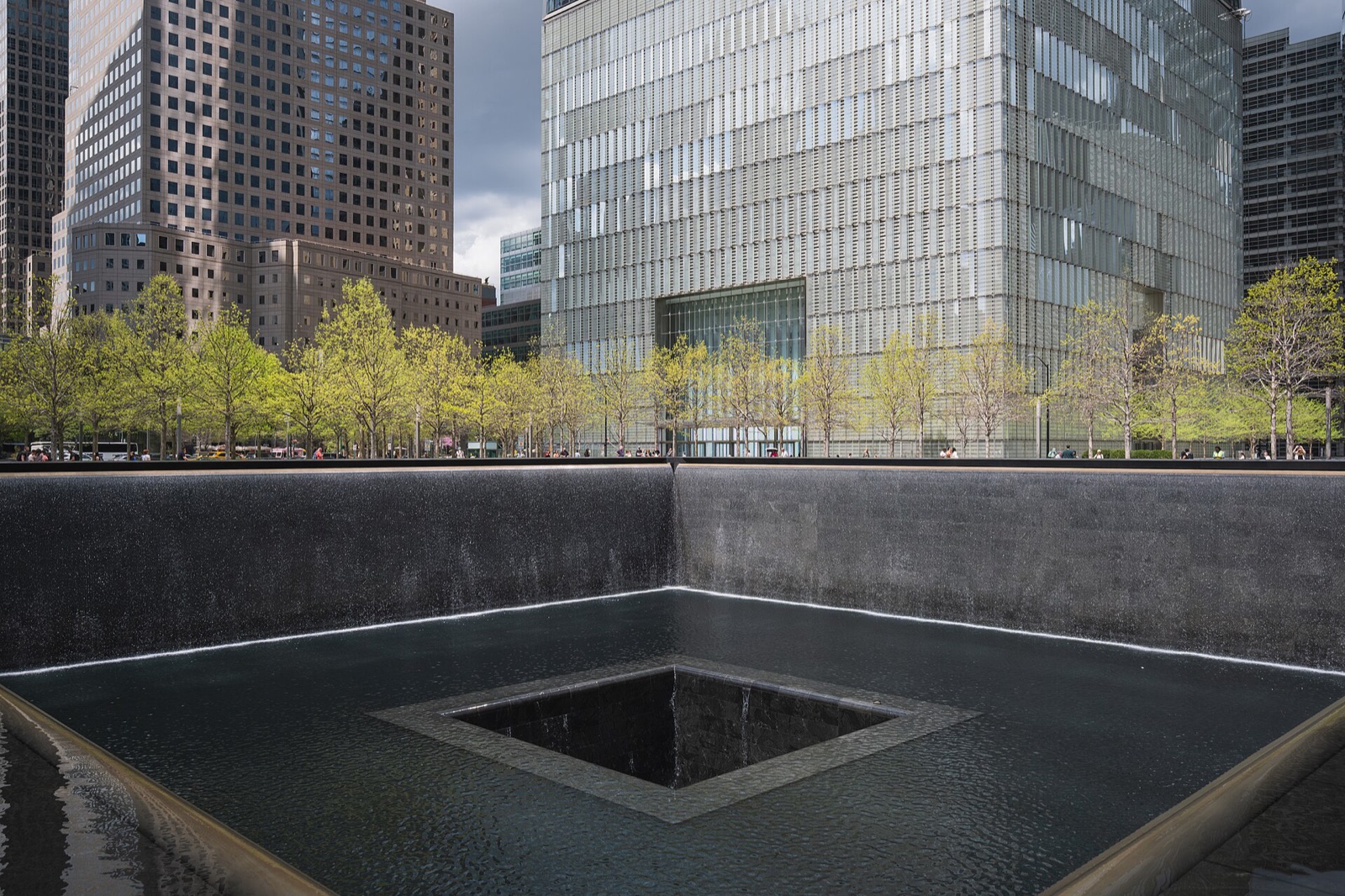 The north pool of the 9/11 Memorial among its oak trees, with the towers of the World Trade Center above.