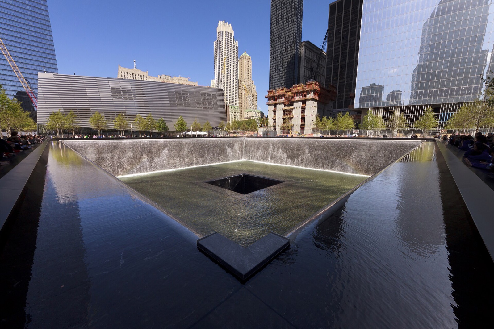 The south pool of the 9/11 Memorial, water falling into a square void, with One World Trade Center and the museum's pavilion behind it.