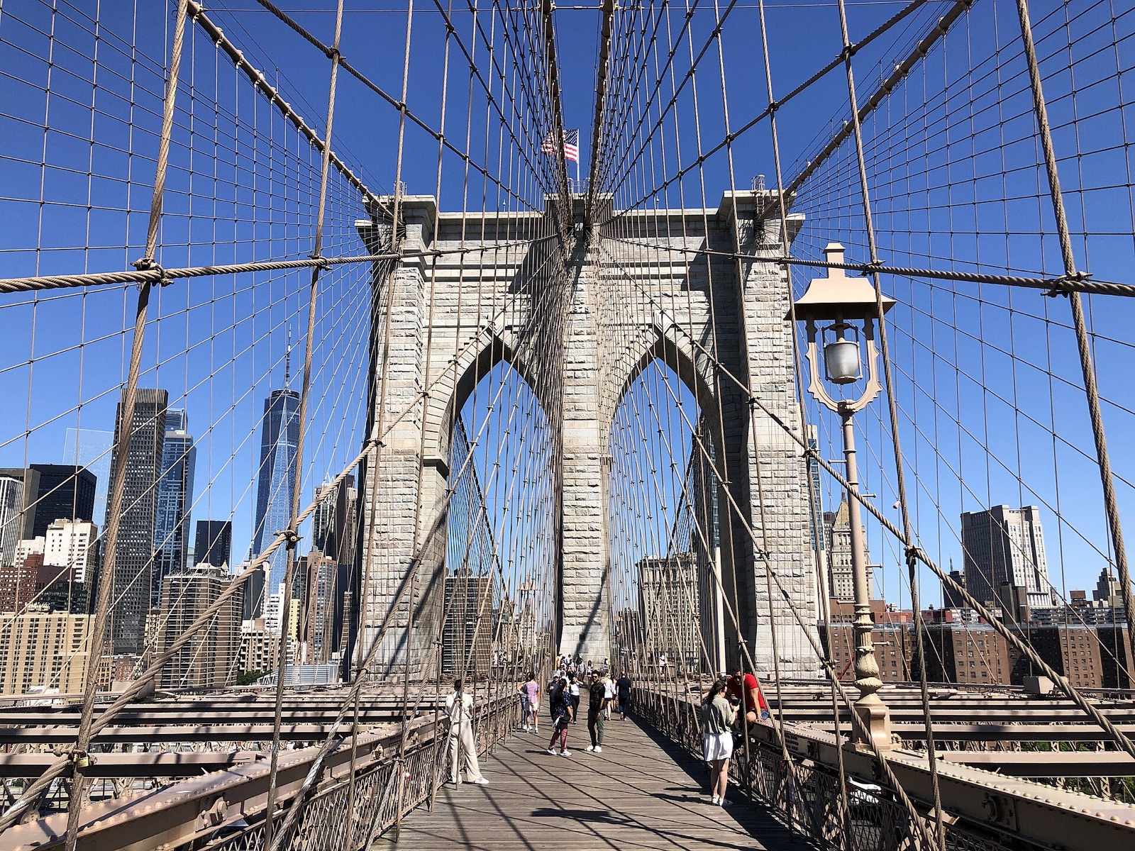 The walkway of the Brooklyn Bridge, leading to a stone tower with two pointed arches, with the skyline of Manhattan beyond.