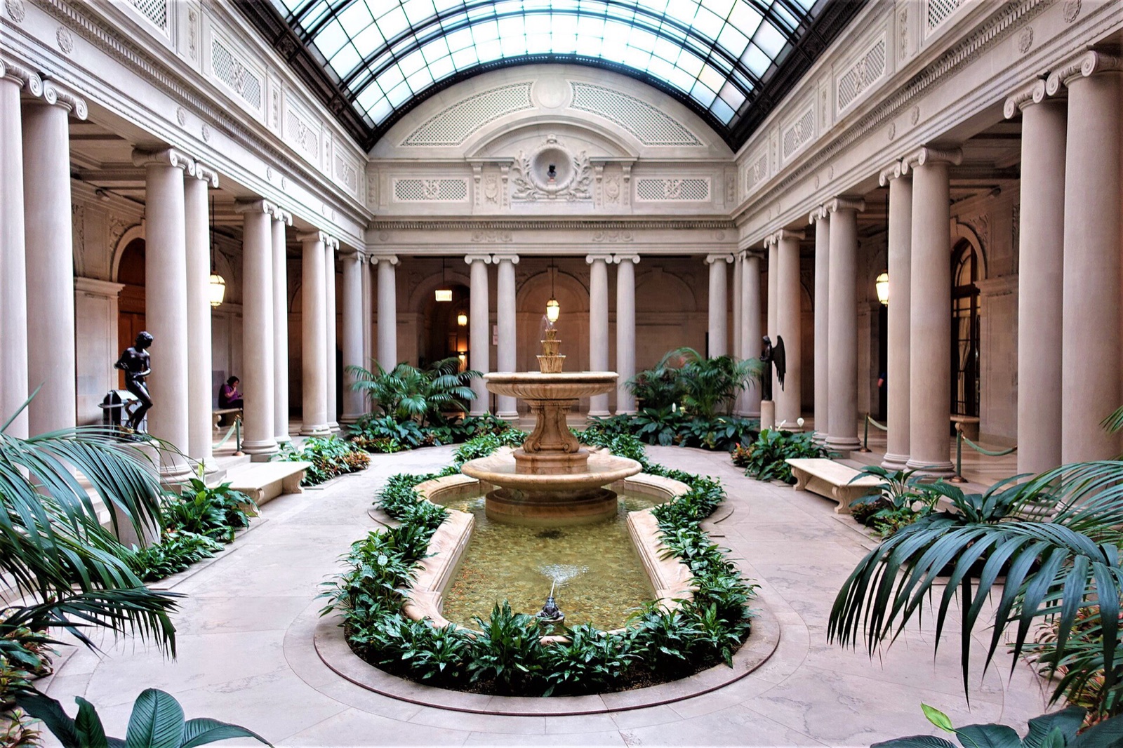 The Garden Court of the Frick Collection, with a fountain and plants under a glass roof ringed by columns.