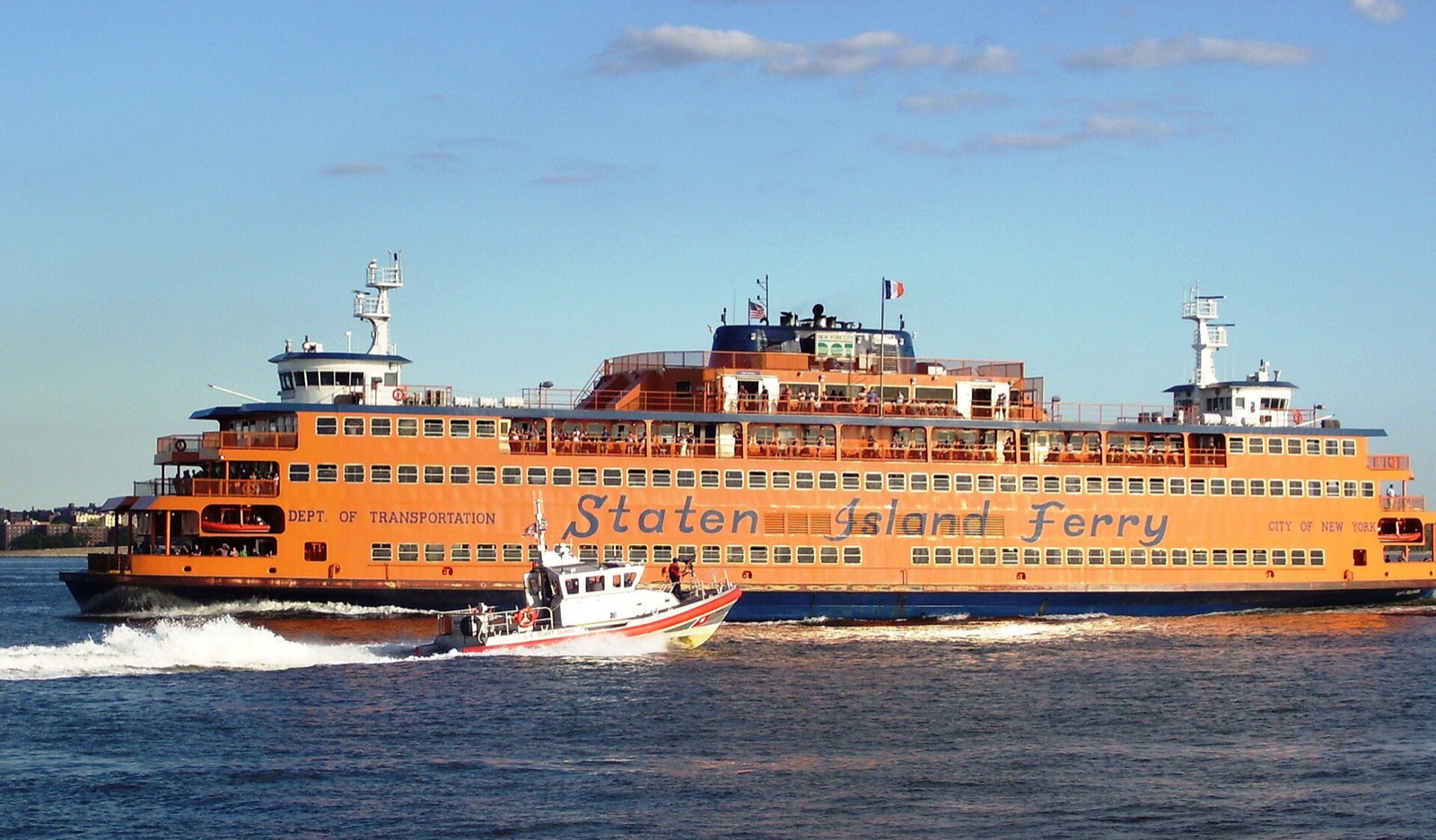 An orange Staten Island Ferry under way in the harbour, with a small patrol boat passing in front of it.