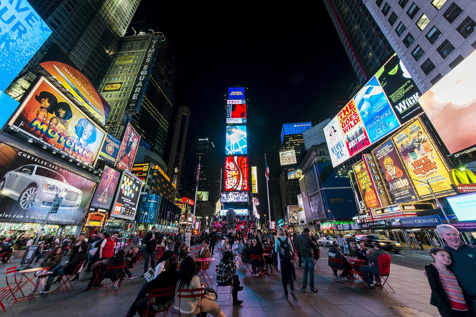 Times Square at night, the screens lit on every building and people sitting at tables in the roadway.