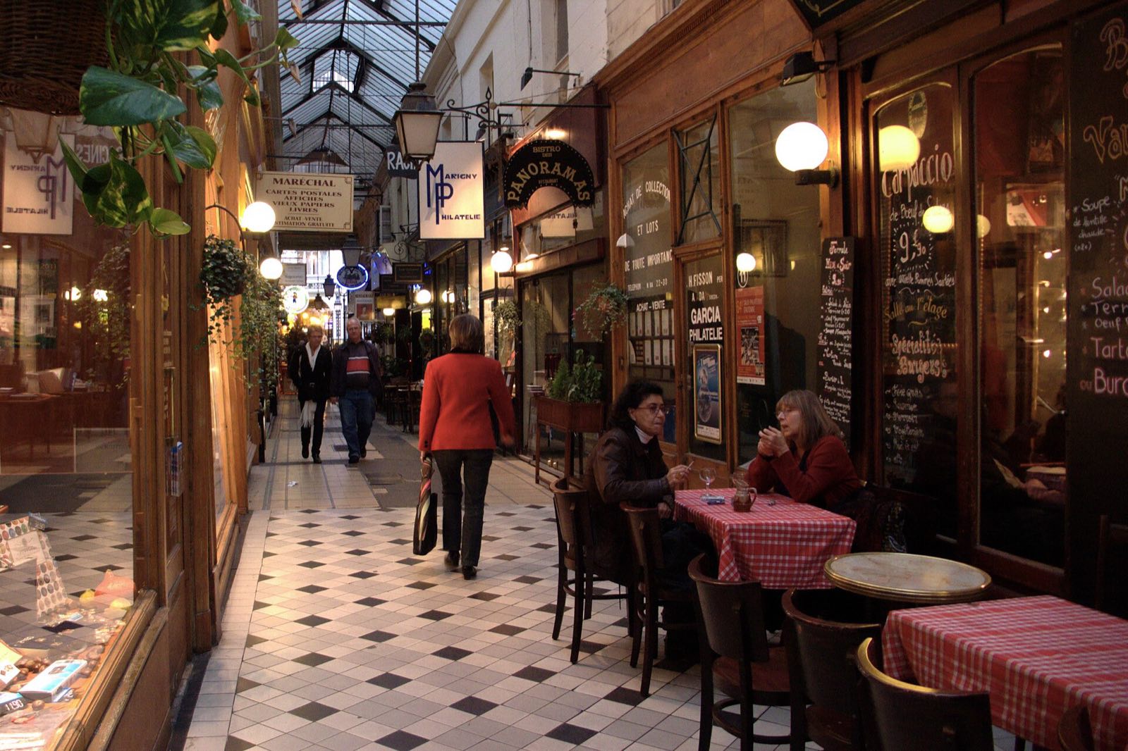The Passage des Panoramas, a narrow arcade with a chequered floor, café tables and shop fronts under a glass roof.