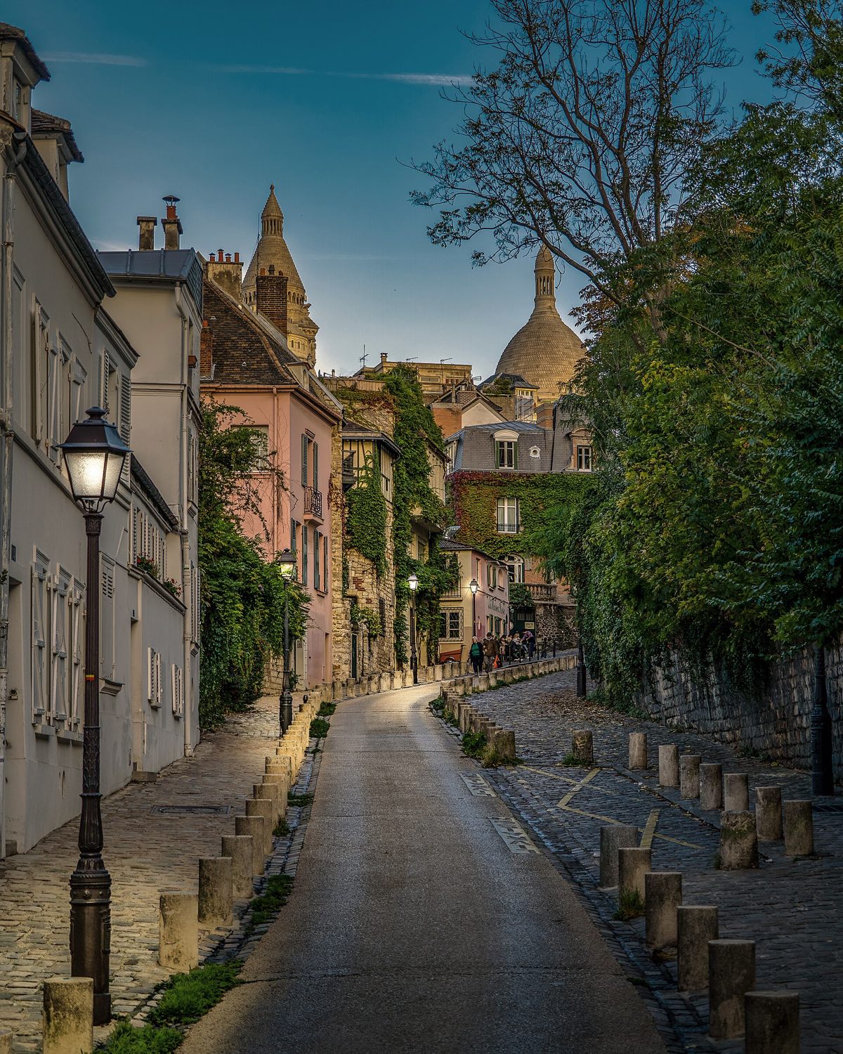 The rue de l'Abreuvoir in Montmartre at dusk, a cobbled lane curving uphill with the dome of the Sacré-Cœur behind.