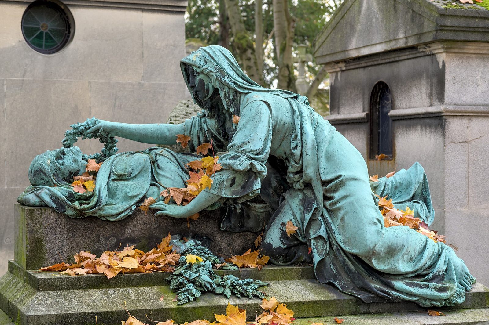 A bronze figure of a mourner lying across a tomb at Père Lachaise, among autumn leaves.