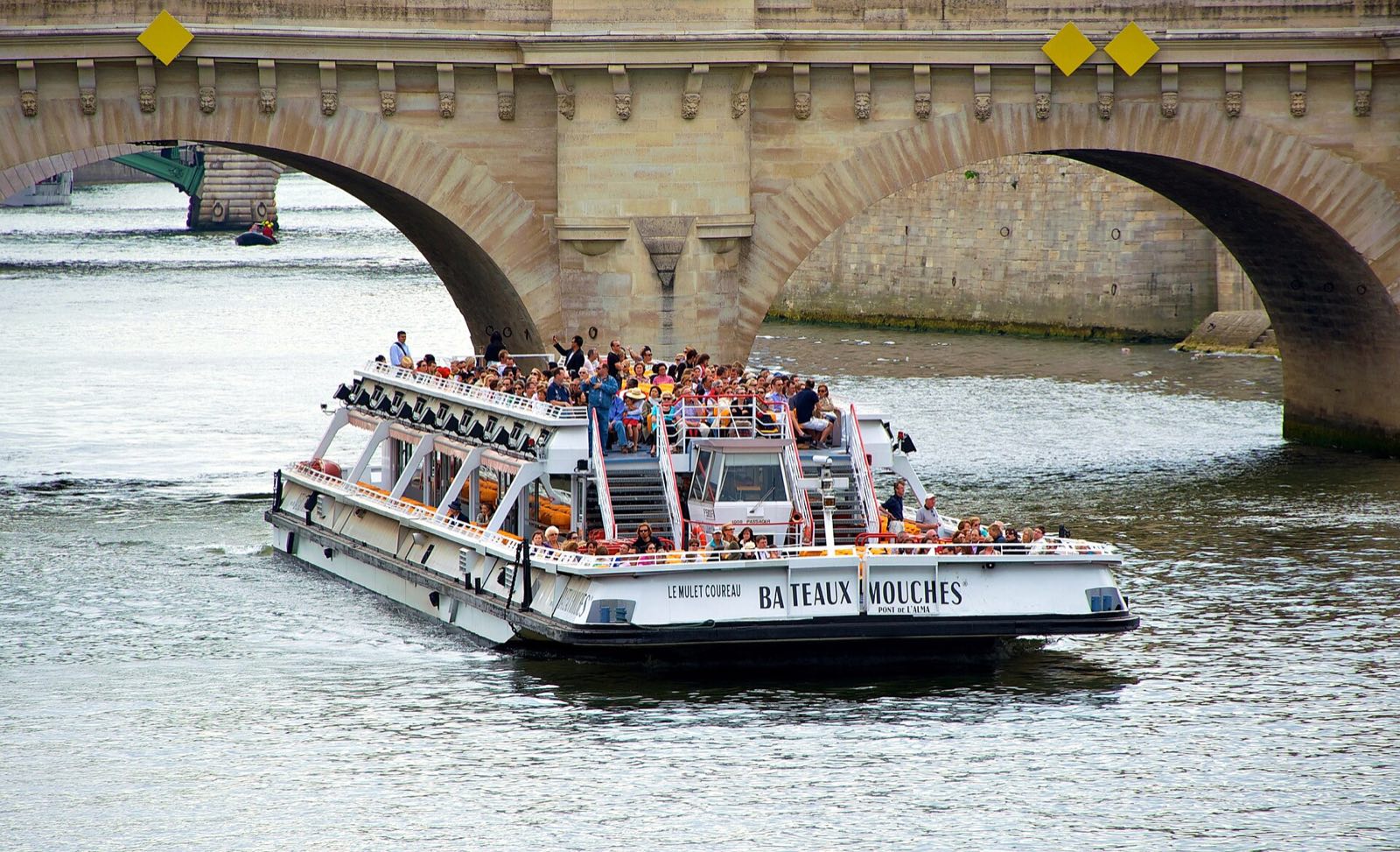 A sightseeing boat full of passengers passing under a stone bridge on the Seine.